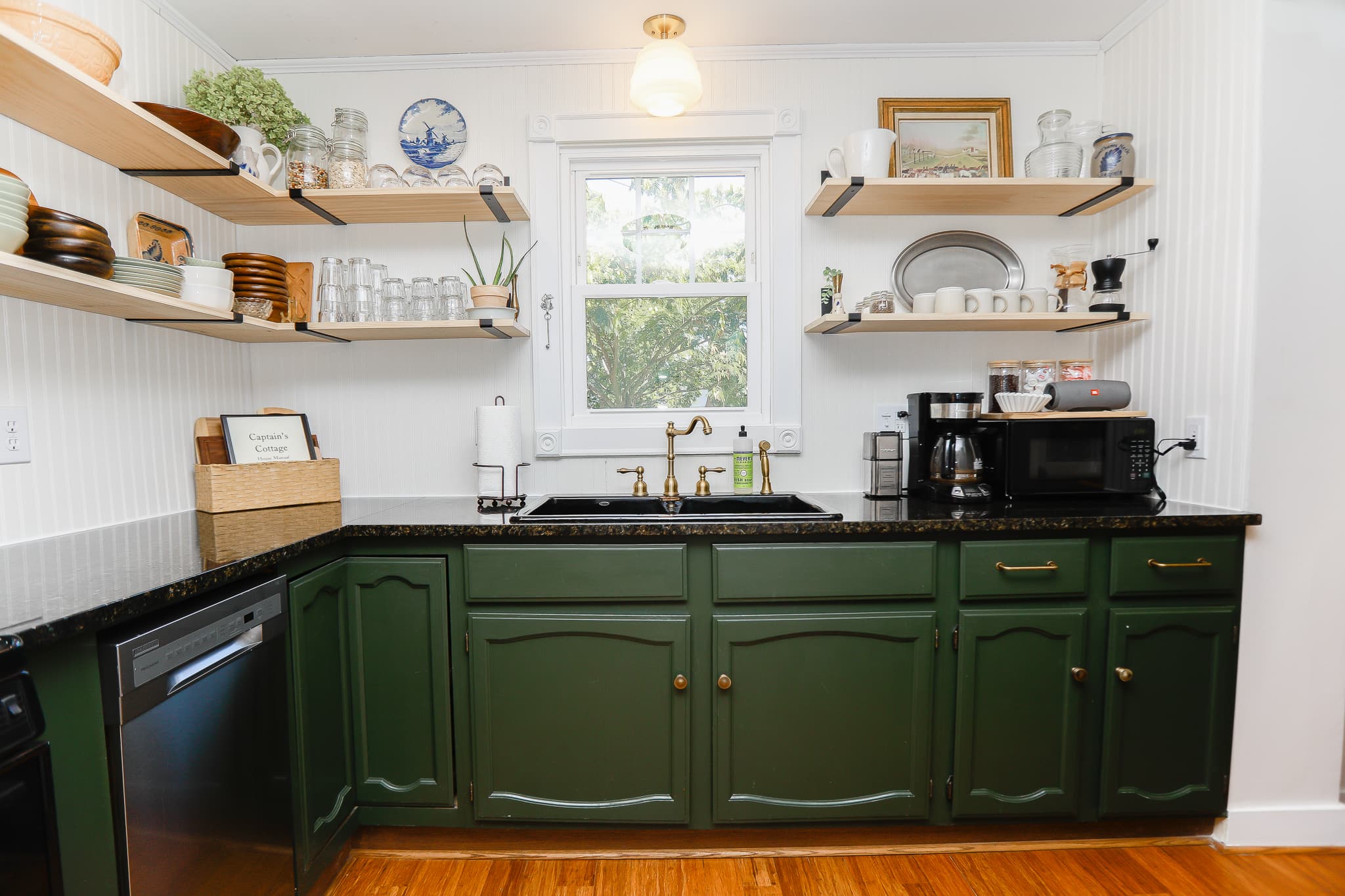 Kitchen at Captain's Cottage — deep-green cabinets, open shelving, brass faucet, and the house manual on the counter.