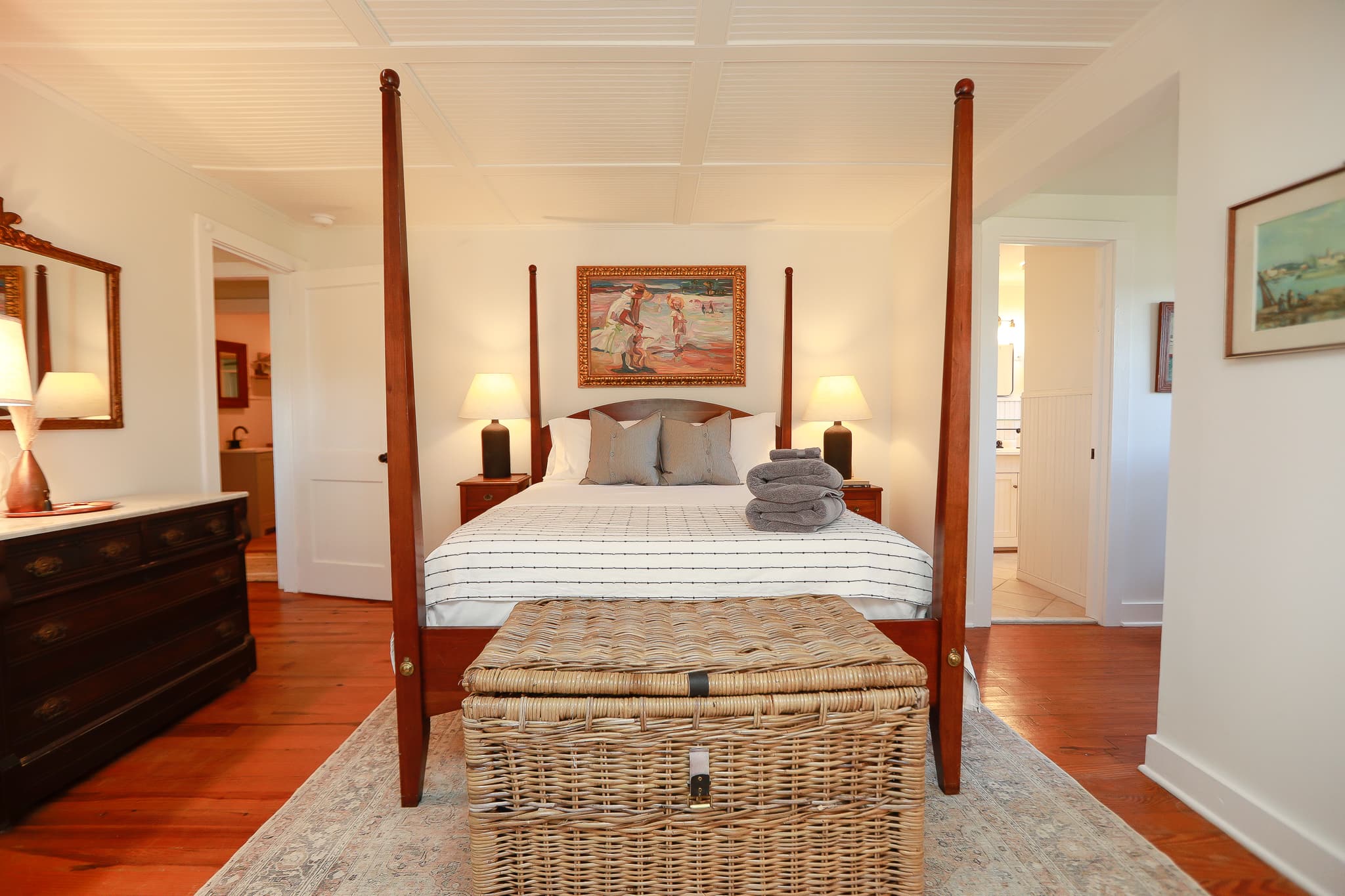 Master bedroom at Captain's Cottage with a mahogany four-poster bed, wicker trunk, and a framed beach painting above the headboard.